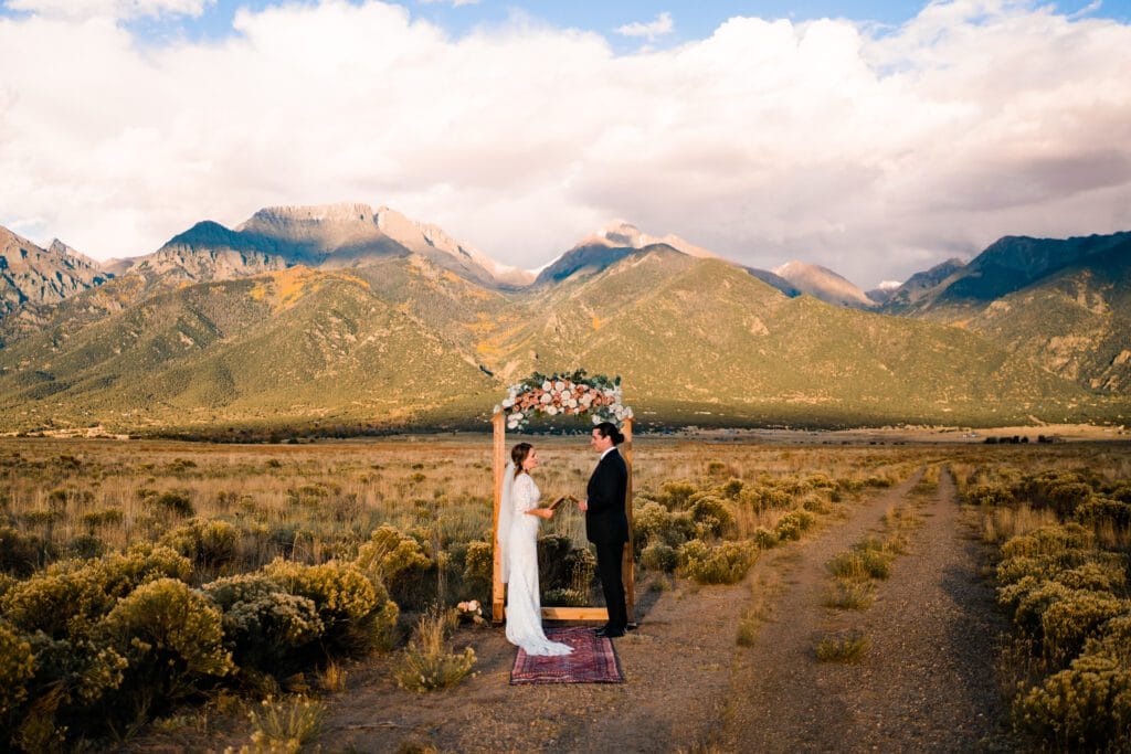 A bride and groom stand under a floral wedding arch on a rug in a field, with mountains and cloudy sky in the background, during an outdoor ceremony.