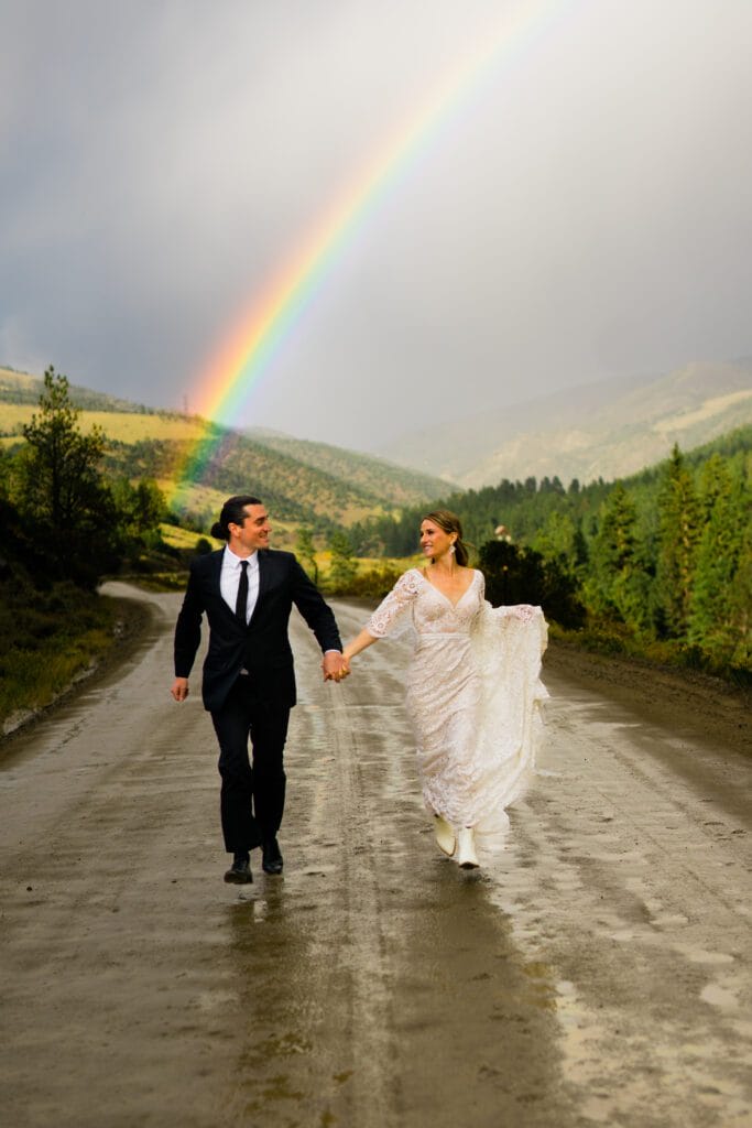 A bride and groom walk hand in hand down a wet country road, surrounded by green trees and hills, with a vibrant rainbow arching across the cloudy sky behind them.