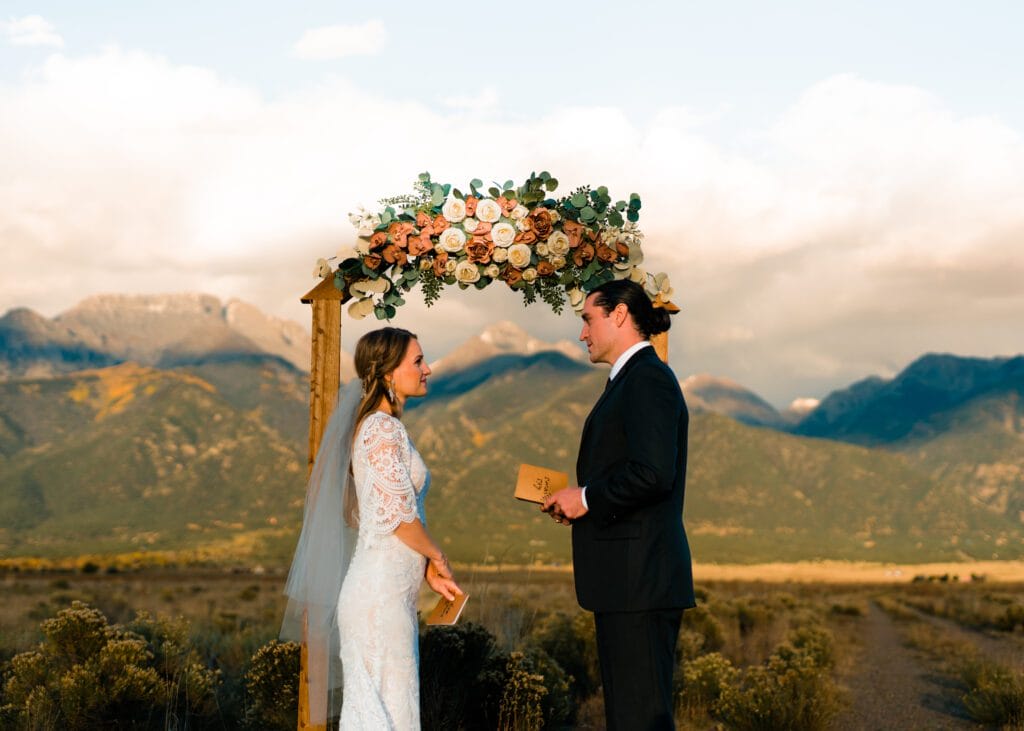 A bride and groom stand under a floral arch in a scenic outdoor setting with mountains in the background, exchanging vows and holding cards, both dressed formally.