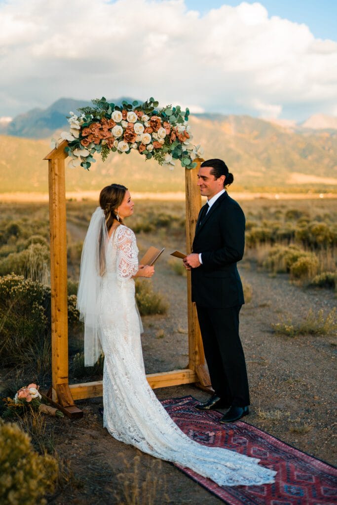 A bride and groom stand facing each other under a wooden arch with flowers, exchanging vows outdoors with mountains and fields in the background. The bride wears a lace dress and veil; the groom is in a dark suit.