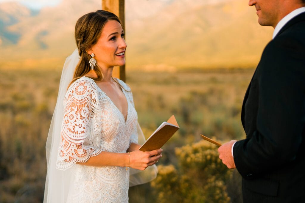 A bride in a lace dress and veil holds a notebook and smiles at a man in a suit during an outdoor wedding ceremony with mountains in the background.