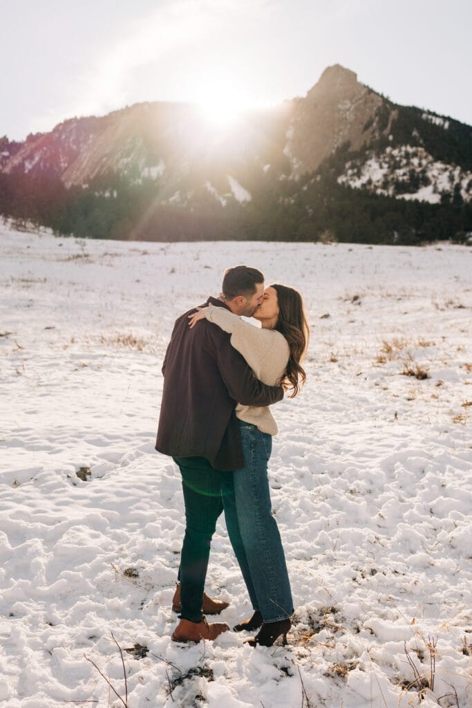 A couple embraces and kisses in a snowy field with mountains in the background. The sun shines brightly over the peaks, casting a warm glow on the winter landscape.