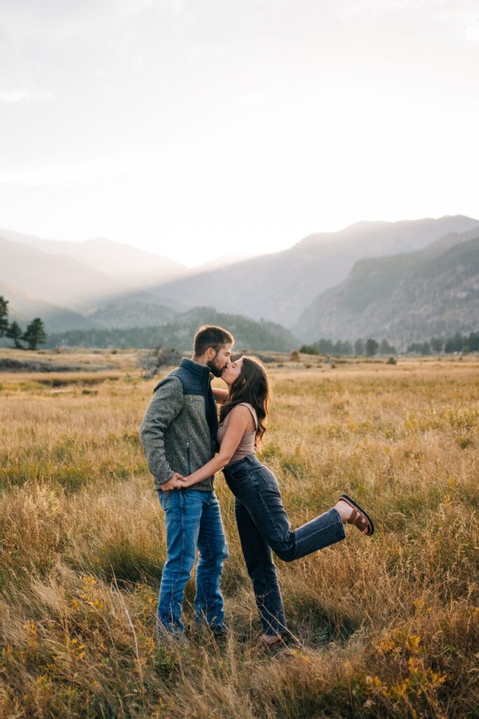 A couple kisses in a golden field at sunset, with mountains in the background. The woman stands on one leg, playfully lifting her other leg behind her as she leans into the kiss.