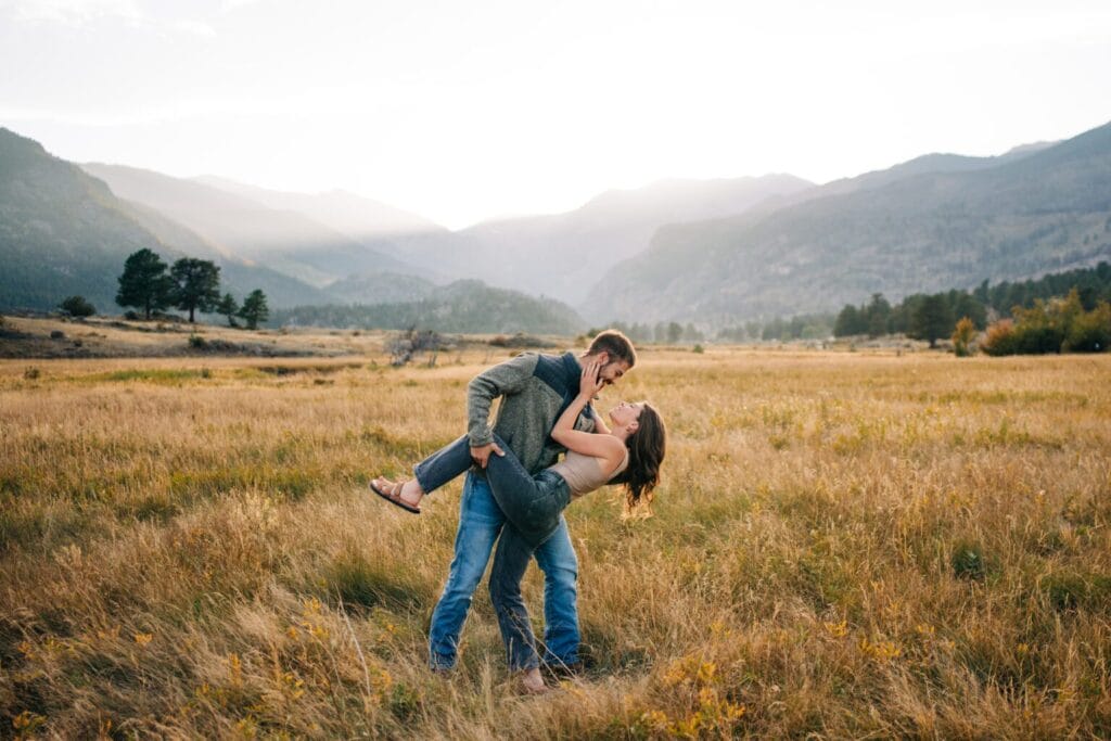 A man lifts and dips a smiling woman in a grassy field at sunset, with mountains and trees in the background under a softly lit sky.