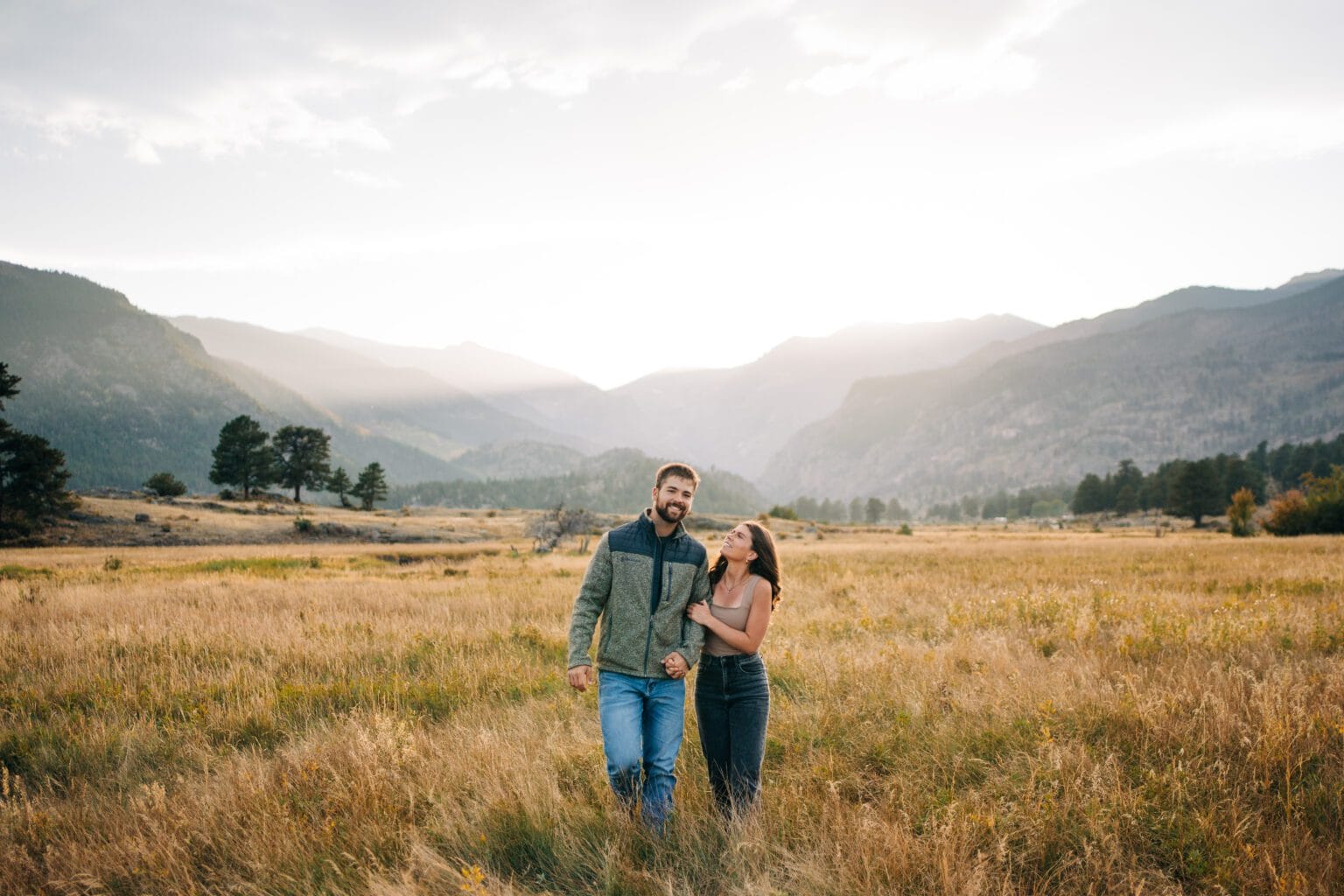 A smiling couple walks hand in hand through a golden grassy field with mountains and trees in the background under a bright, cloudy sky.