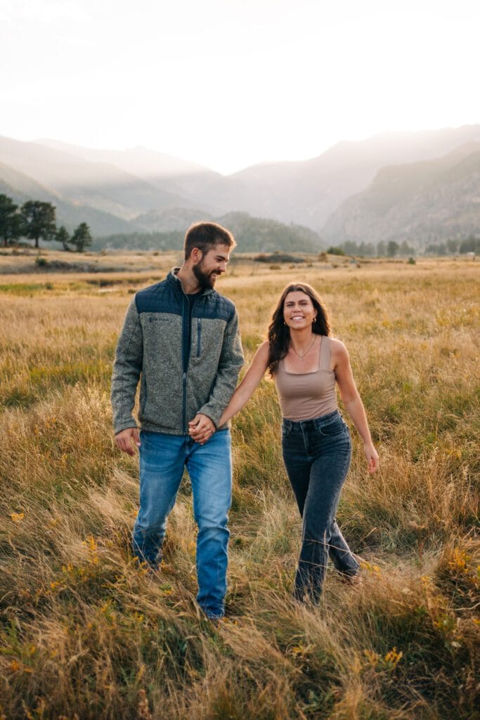 A couple walks hand-in-hand through a grassy field at sunset, smiling and enjoying the scenic mountain landscape in the background.