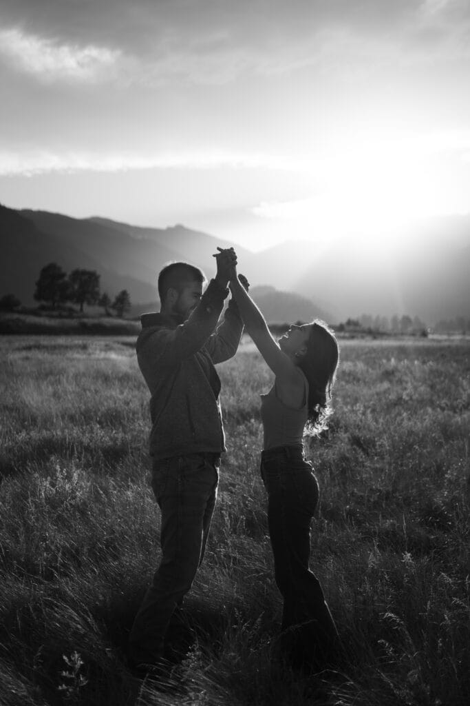 A couple stands in a grassy field at sunset, holding hands and facing each other, with mountains visible in the background and sunlight streaming through the clouds.