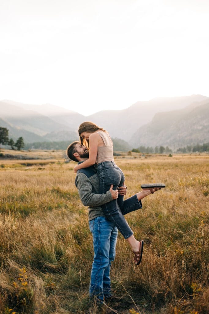 A couple embraces in a grassy field with mountains in the background. The man lifts the woman as she leans in to kiss him, both smiling and dressed casually. The scene is warm and outdoors at sunset.
