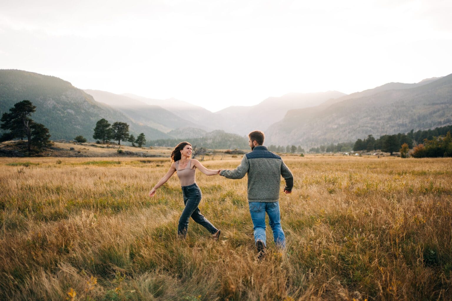A woman and man holding hands, running through a grassy field with mountains in the background during sunset. They appear happy and playful, surrounded by nature.