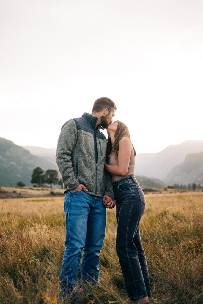 A couple stands in a grassy field at sunset, holding hands and sharing a kiss. Mountains and trees are visible in the background, and the sky is softly lit.