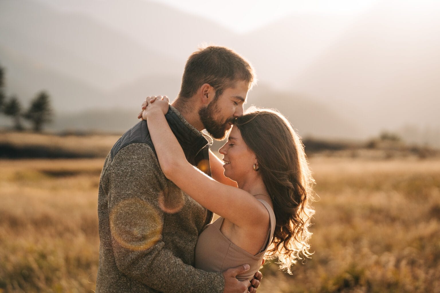 A couple embraces in a sunlit meadow with mountains in the background. The man gently kisses the womans forehead while she smiles, both surrounded by warm, golden light and lens flare.