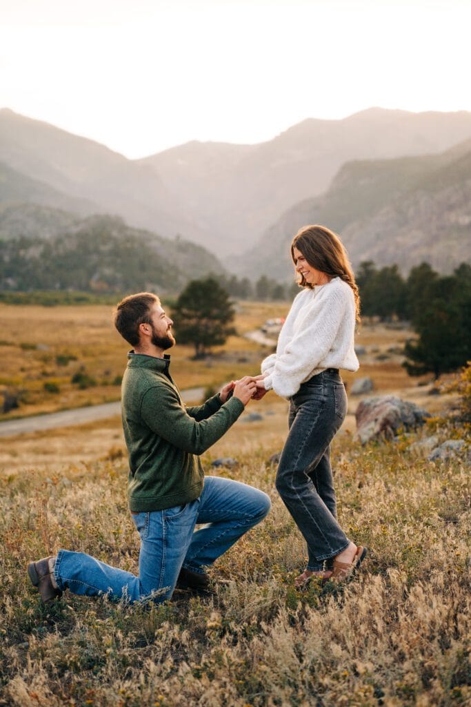 A man kneels on one knee, holding a woman’s hands and proposing to her in a grassy field with mountains and trees in the background at sunset.