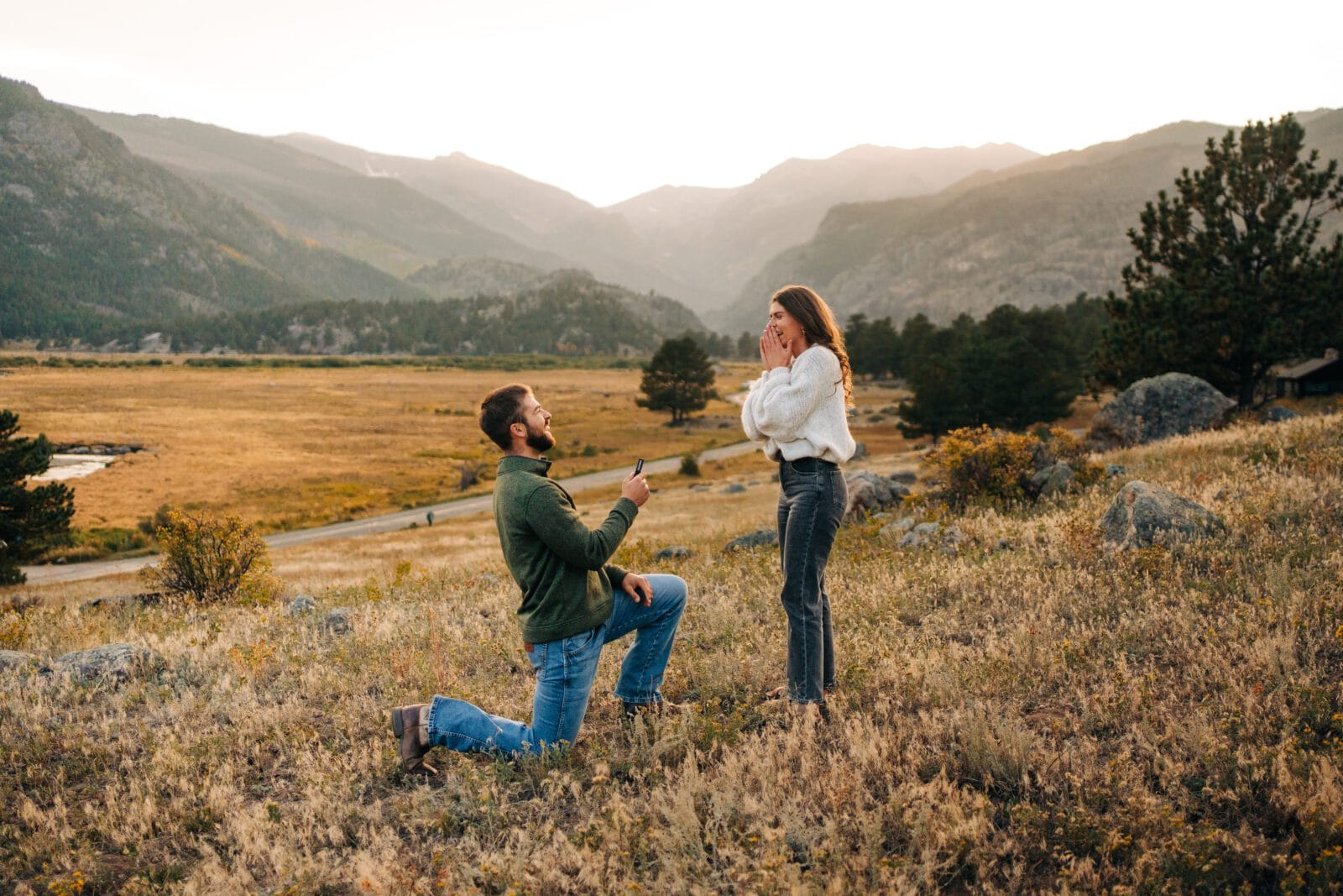 A man kneels on one knee holding a ring box, proposing to a surprised woman in a grassy field with mountains in the background at sunset.
