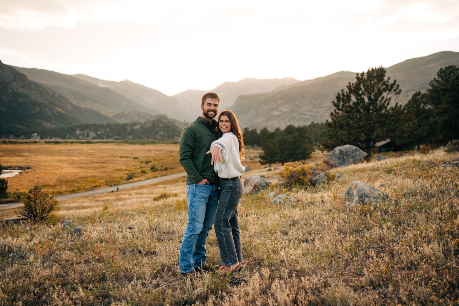 A couple stands on a grassy hillside at sunset, smiling at the camera. The woman extends her hand forward, showing off a ring. Mountains and a valley are visible in the background.