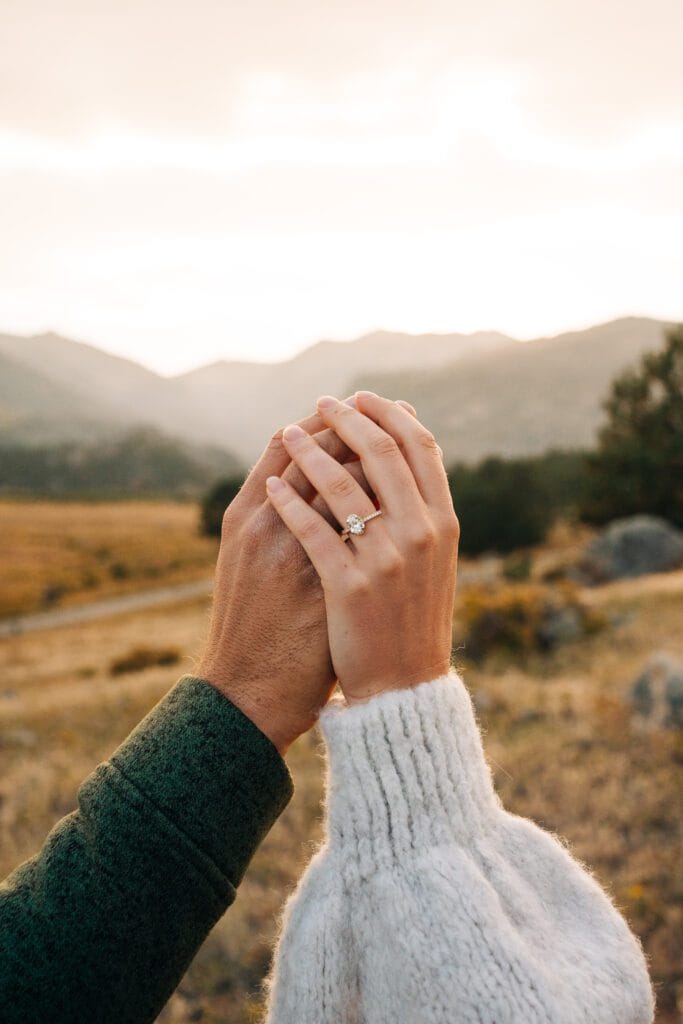 Two people hold hands outdoors with a mountain landscape in the background. One hand wears a diamond engagement ring, and both wear cozy sweaters. The scene is warmly lit by the setting sun.