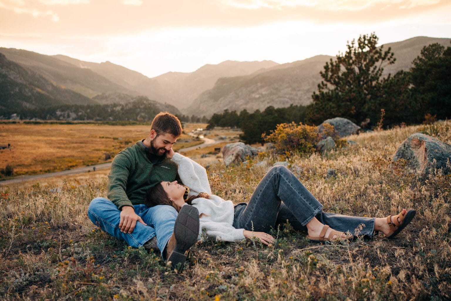 A couple relaxes on a grassy hillside at sunset, with mountains in the background. The man sits upright while the woman lies back, resting her head on his lap, both smiling and enjoying the peaceful outdoor scene.
