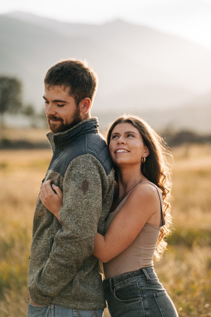 A woman smiles and hugs a man from behind in a sunlit field, with mountains blurred in the background. The man looks down and smiles softly, while the woman gazes up at him.