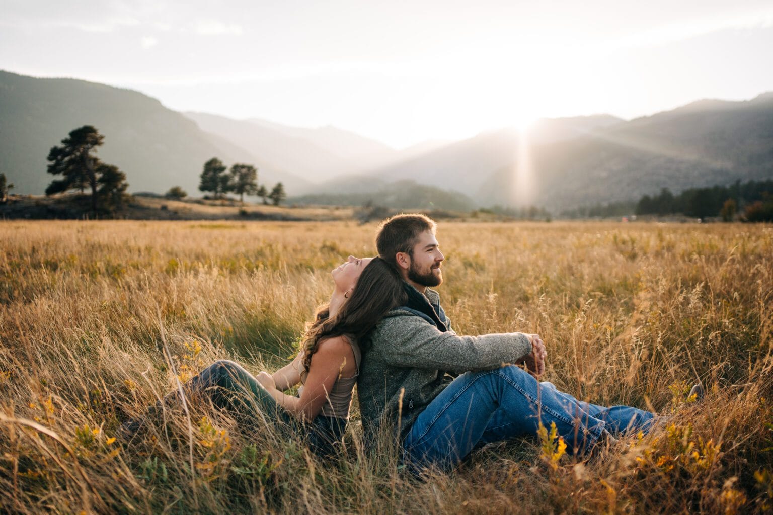 A couple sits back-to-back in a grassy field at sunset, surrounded by mountains and trees, with the sun shining through the clouds in the background. The scene feels peaceful and intimate.