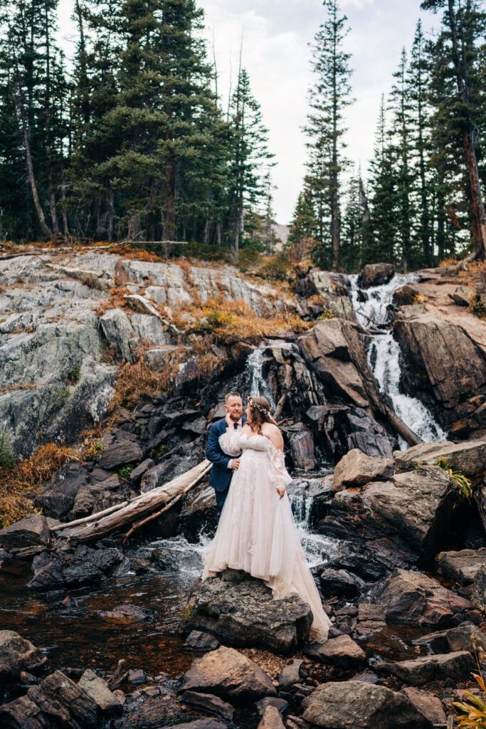 A bride and groom stand on rocks in front of a small waterfall and forest, embracing each other in wedding attire. Tall pine trees and rugged terrain surround them, creating a natural, scenic backdrop.