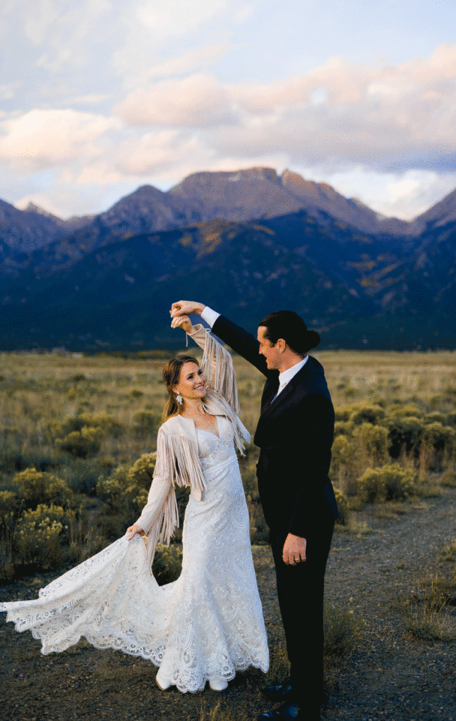 A couple dressed in wedding attire dances outdoors in front of scenic mountains and an open field, the woman smiling as she twirls in her lace dress while the man holds her hand.