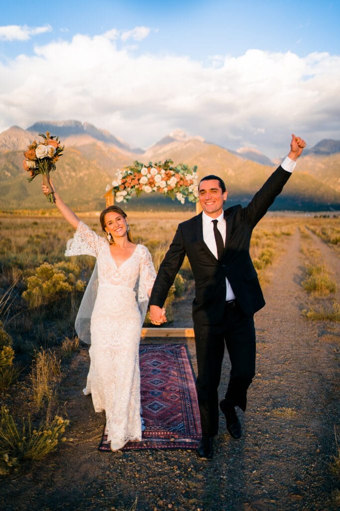 A bride in a white lace dress and a groom in a black suit walk hand-in-hand down an outdoor aisle, both smiling and raising their arms in celebration, with mountains and a floral arch in the background.
