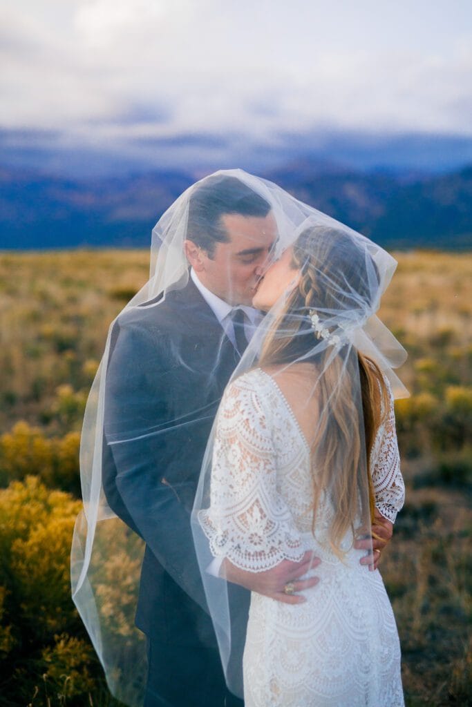 A bride and groom kiss outdoors in a field, wrapped together under a sheer veil. The bride wears a white lace dress, and the groom is in a dark suit. Mountains and a cloudy sky are visible in the background.