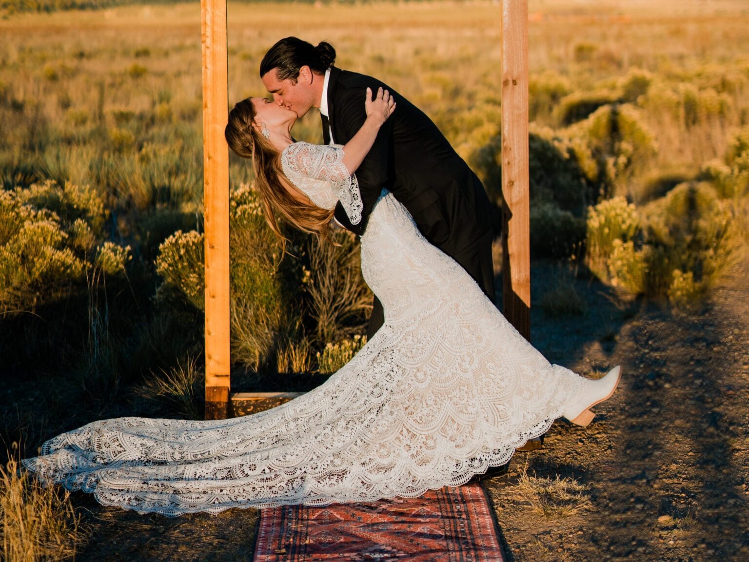A bride in a long, lace wedding dress is dipped backward in a kiss by a groom in a black suit, standing outdoors on a rug with golden fields and sunlight in the background.