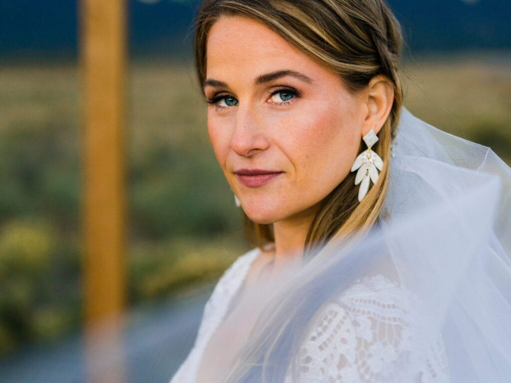 A woman in a white lace wedding dress and veil, with long hair and large floral earrings, looks at the camera with a serene expression. The background is blurred, showing an outdoor setting.