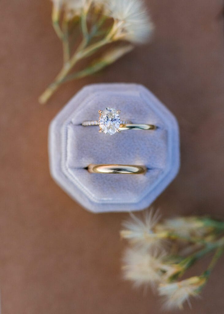 A gold wedding band and a diamond solitaire engagement ring displayed in a light gray velvet ring box, surrounded by soft, out-of-focus dried flowers on a brown surface.
