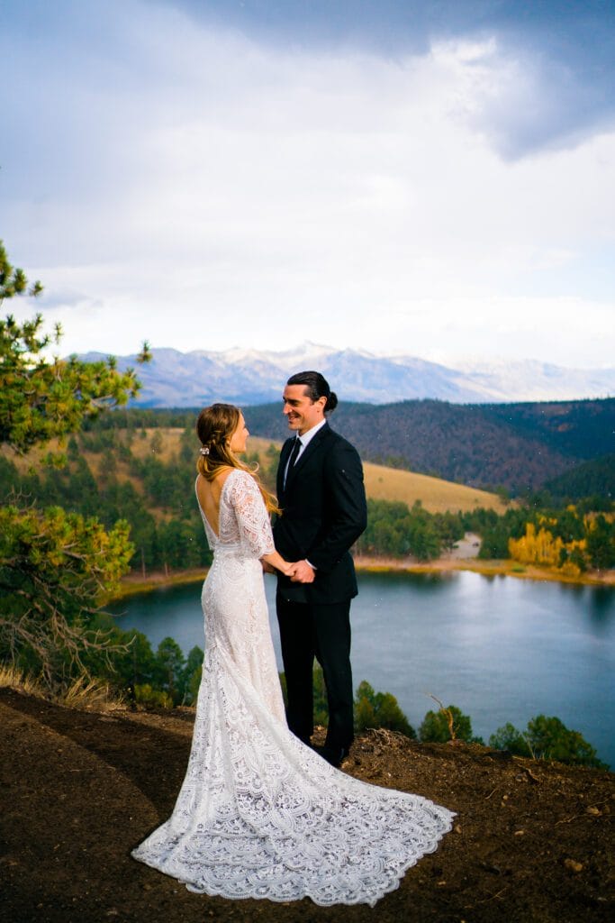 A bride and groom stand holding hands on a hill overlooking a scenic lake and mountains, with trees and cloudy skies in the background. The bride wears a white lace gown, and the groom is in a black suit.