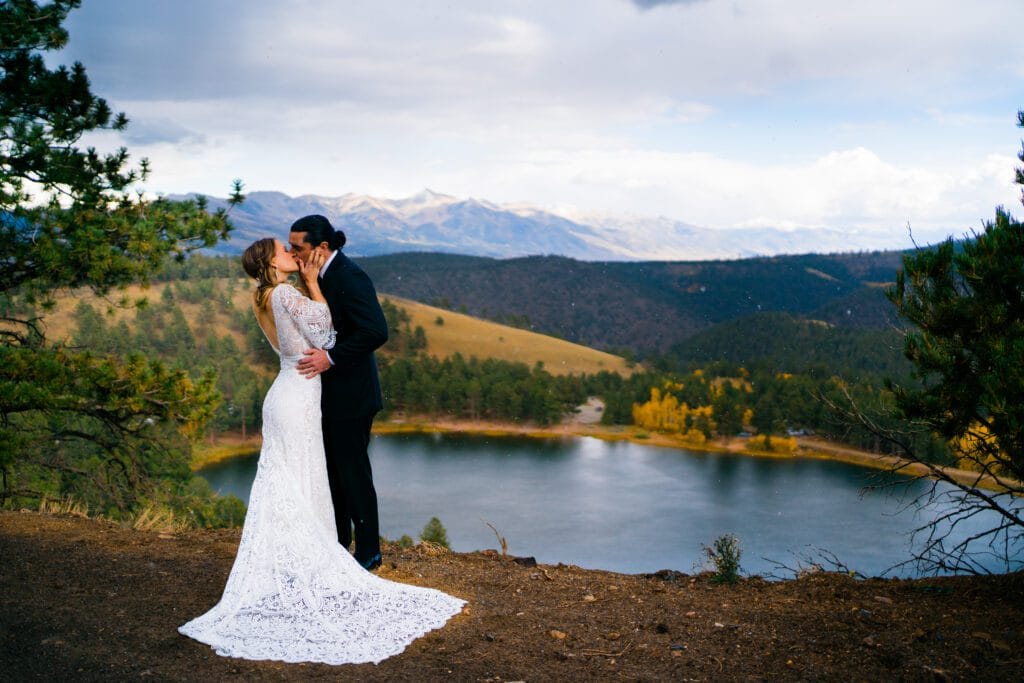 A bride and groom kiss on a hilltop overlooking a scenic lake, surrounded by trees and mountains under a partly cloudy sky.