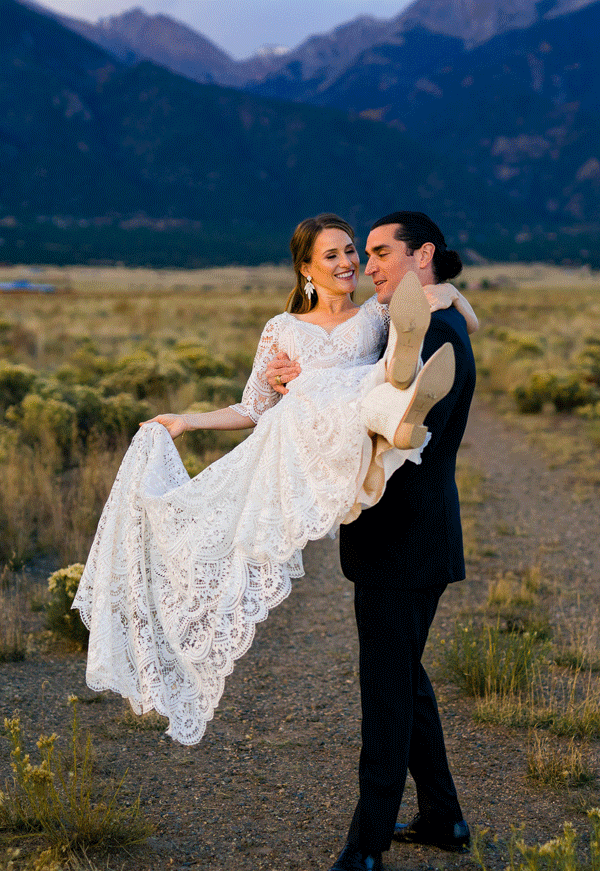 A groom in a black suit carries his bride, who is wearing a white lace wedding dress, across a field with mountains in the background. Both are smiling and looking at each other lovingly.