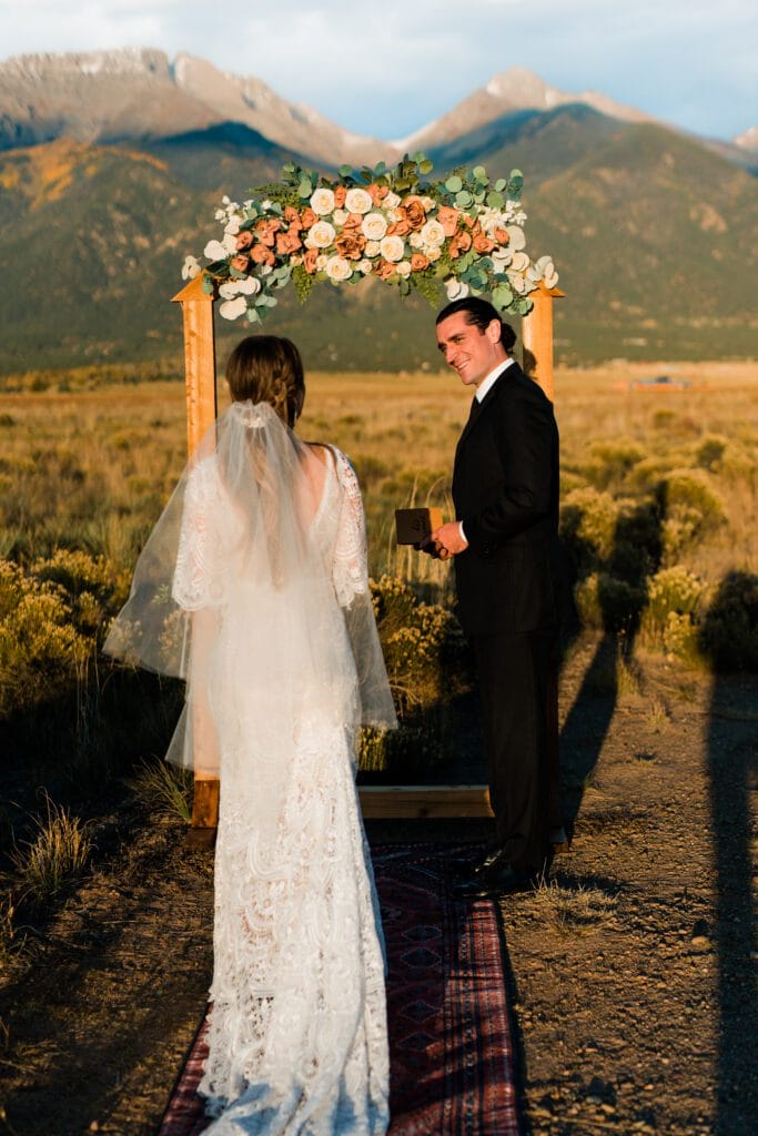 A bride in a lace gown and veil stands facing a groom in a black suit under a floral arch outdoors, with mountains and fields in the background, during a wedding ceremony at sunset.