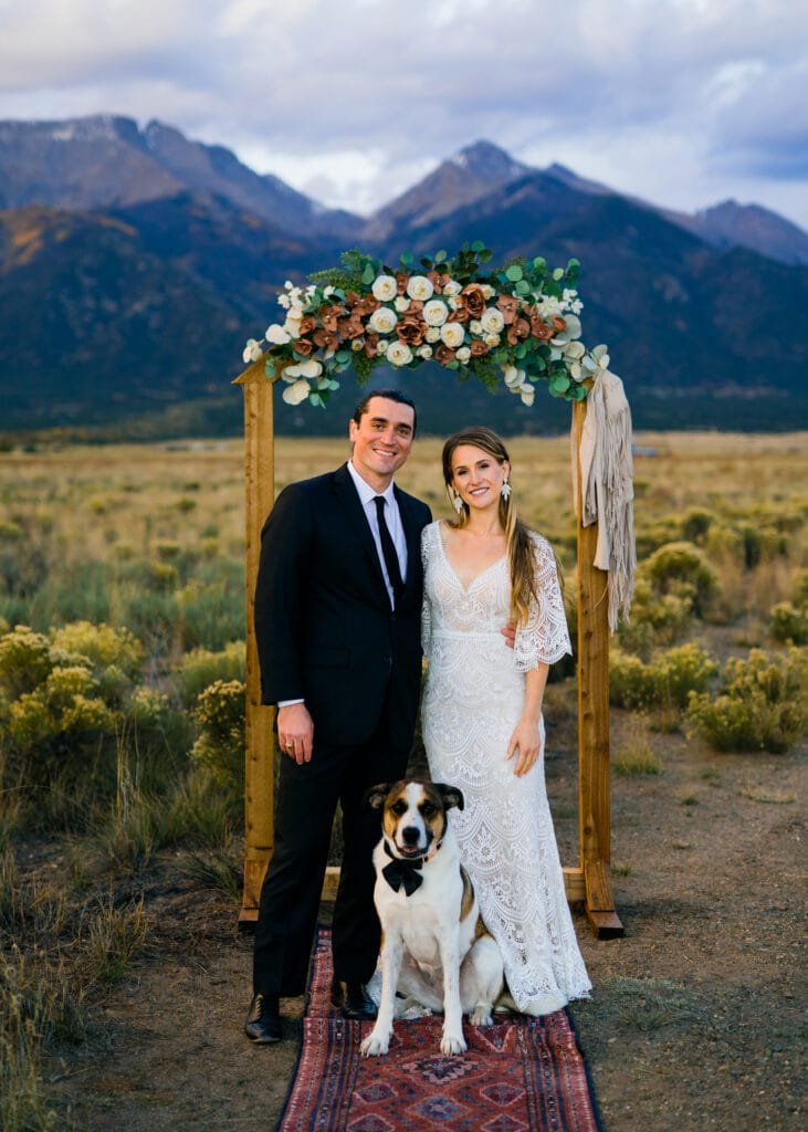 A bride and groom stand smiling under a floral wedding arch in a scenic outdoor setting with mountains in the background. A dog sits in front of them on a patterned rug.