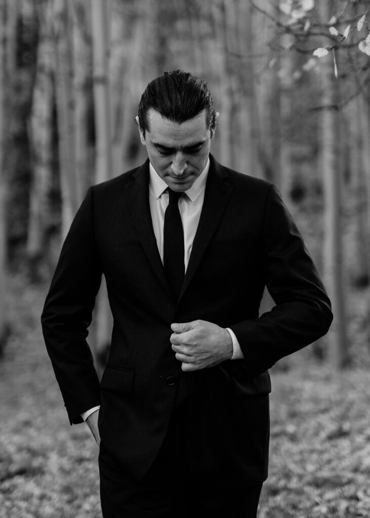 A man in a dark suit and tie stands outdoors, looking down as he buttons his jacket with one hand. The background is blurred with tall trees, and the image is in black and white.