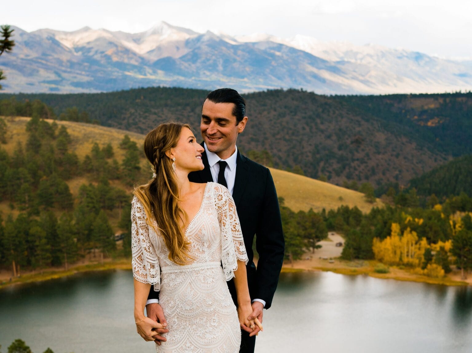 A bride and groom stand close together, smiling at each other, with a scenic mountain landscape, forest, and lake in the background. The bride wears a lace dress and the groom is in a black suit.