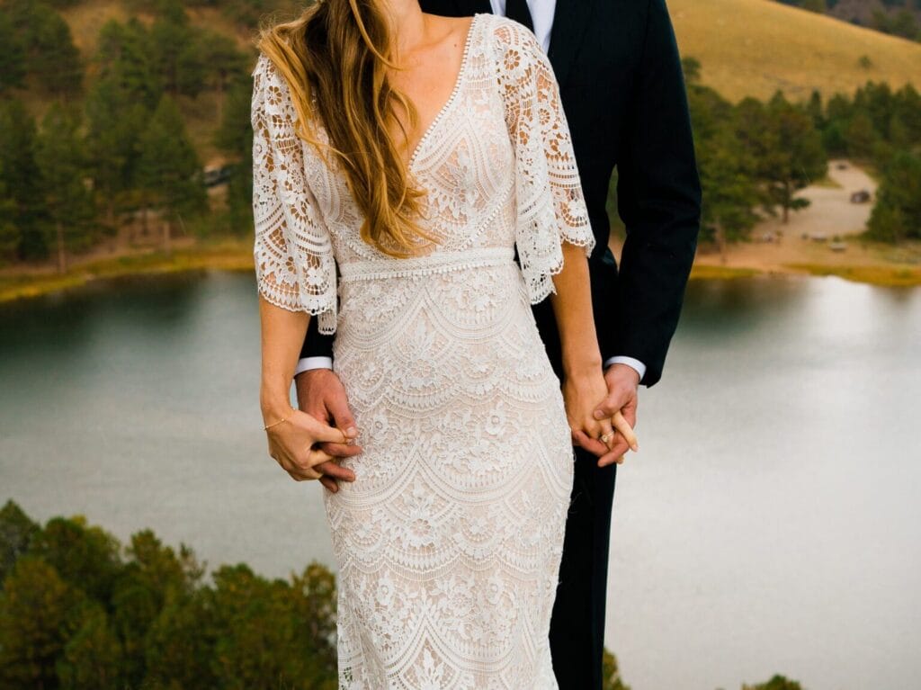A couple dressed in wedding attire stands outdoors near a lake, holding hands. The bride wears a lace dress with elbow-length sleeves, and the groom wears a dark suit. The background features trees and rolling hills.