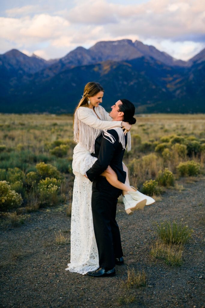 A man in a black suit lifts a woman in a white lace dress and fringe jacket. They smile at each other in a field, with tall mountains and a cloudy sky in the background.