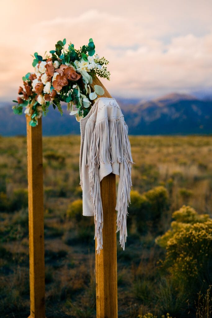 A wooden wedding arch decorated with a floral arrangement and a white fringed jacket, set in an open field with mountains in the background at sunset.
