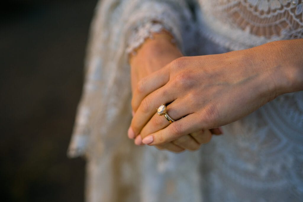A close-up of a persons hands wearing a lace dress, with one hand gently clasping the other. The person is wearing a gold engagement ring with a large stone, stacked with a wedding band.