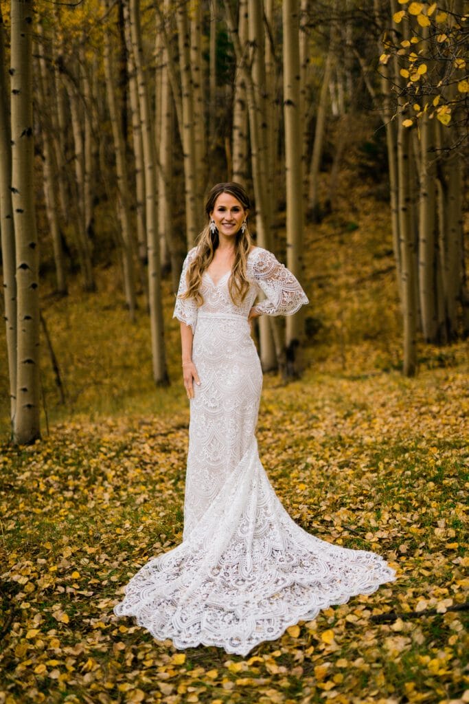 A woman in a long, white lace wedding dress stands smiling in a forest with tall trees and yellow autumn leaves covering the ground.