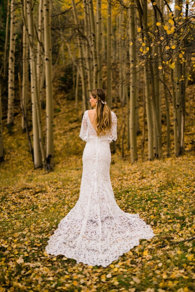 A woman in a white lace wedding dress stands in a forest of tall, slender trees with yellow leaves scattered on the ground, facing away from the camera.