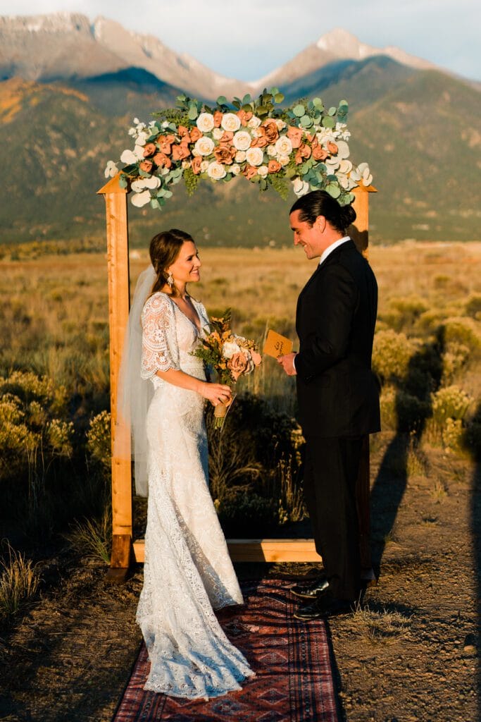 A bride and groom stand under a wooden arch adorned with flowers, exchanging vows outdoors in a field with mountains in the background, bathed in warm sunlight.