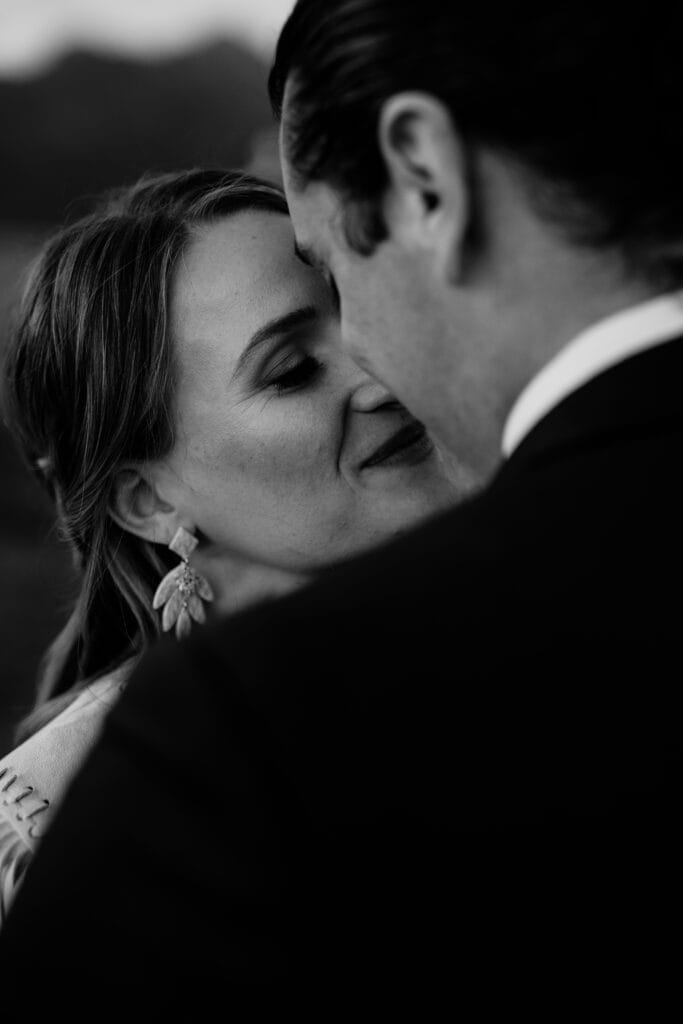 A close-up, black and white photo of a couple embracing intimately. The woman has her eyes closed and a soft smile, wearing leaf-shaped earrings. The man’s face is partially visible, turned toward her.