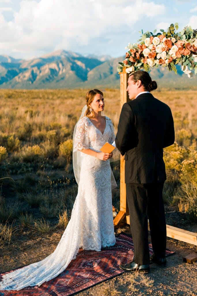A bride in a lace dress and veil holds a booklet and faces a groom in a black suit during an outdoor wedding ceremony, with mountains and greenery in the background. They stand under a floral arch on a patterned rug.