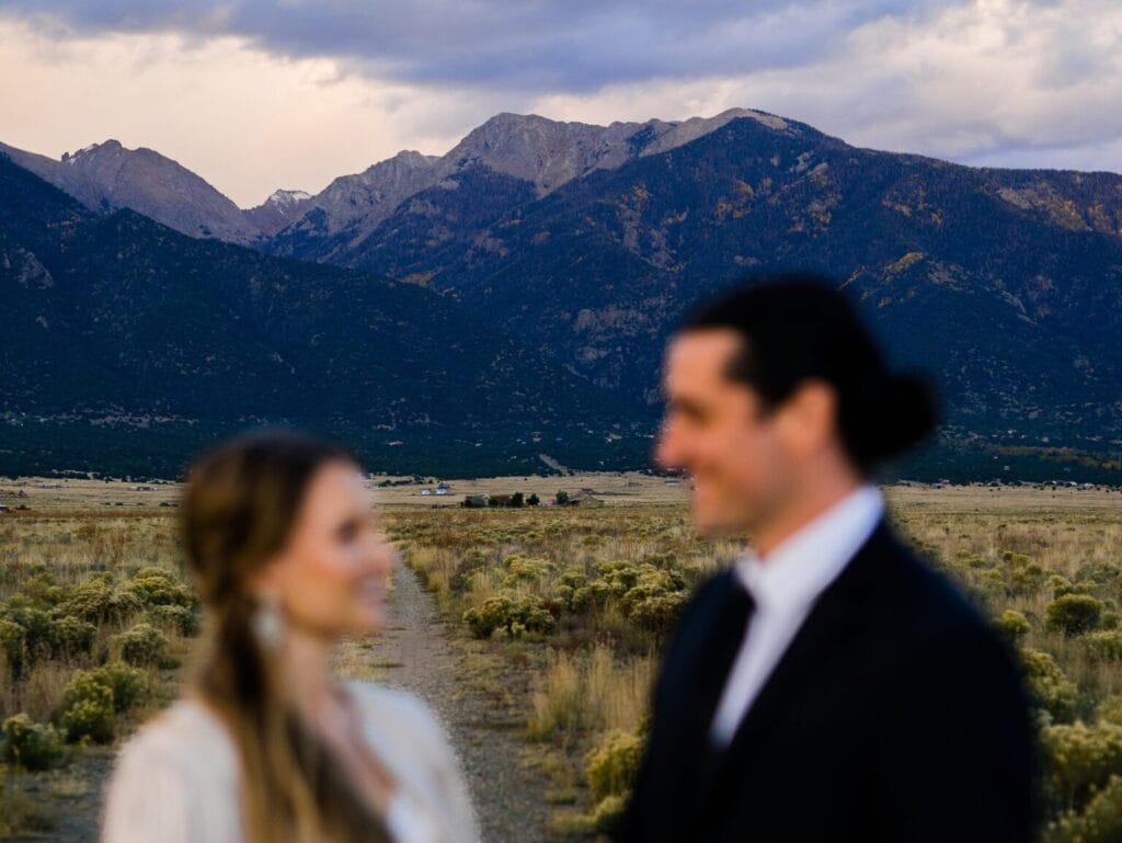 A blurred couple stands in the foreground of a scenic landscape featuring mountains, grassy fields, and a cloudy sky in the background.