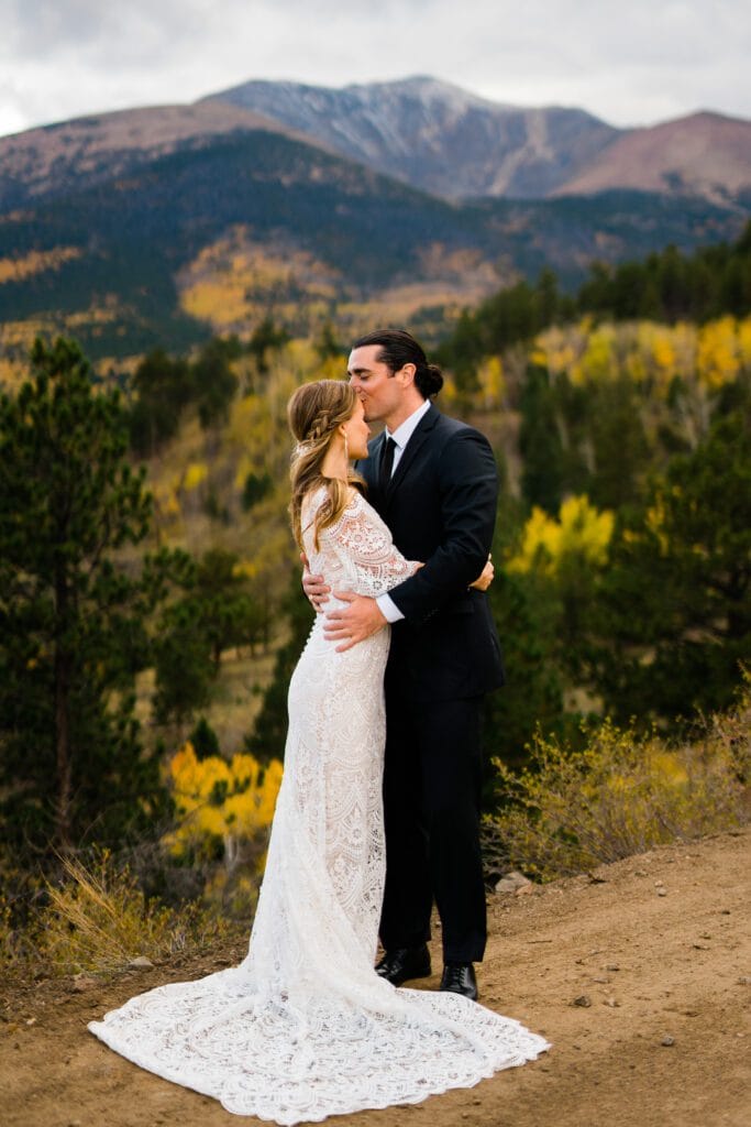 A bride in a white lace dress and a groom in a black suit embrace outdoors on a dirt path with autumn trees and a mountain in the background. The groom kisses the brides forehead.