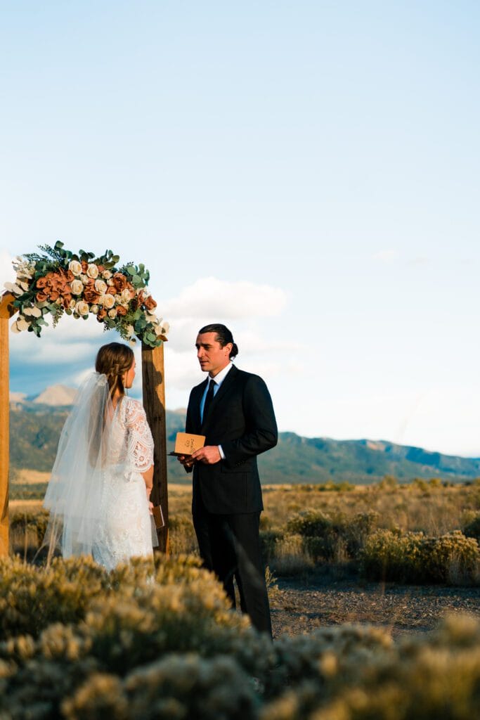 A bride and groom stand outdoors under a floral arch, exchanging vows. The bride wears a white dress and veil, while the groom is in a black suit holding a small book. Mountains and a clear sky are in the background.