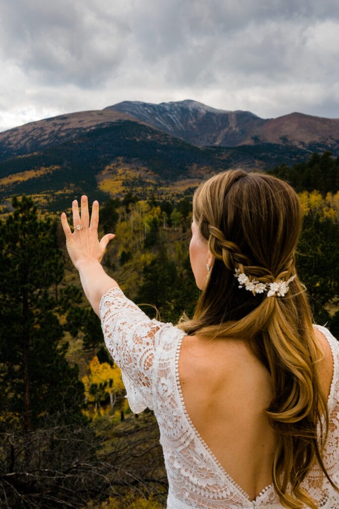 A bride with long, wavy hair and a floral hairpiece raises her hand toward scenic mountains under a cloudy sky, standing among trees with autumn foliage.