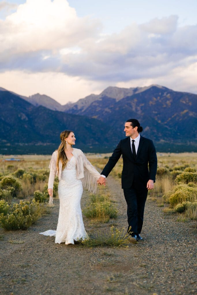 A bride in a white lace dress and a groom in a black suit hold hands while walking outdoors on a gravel path, with mountains and cloudy sky in the background.